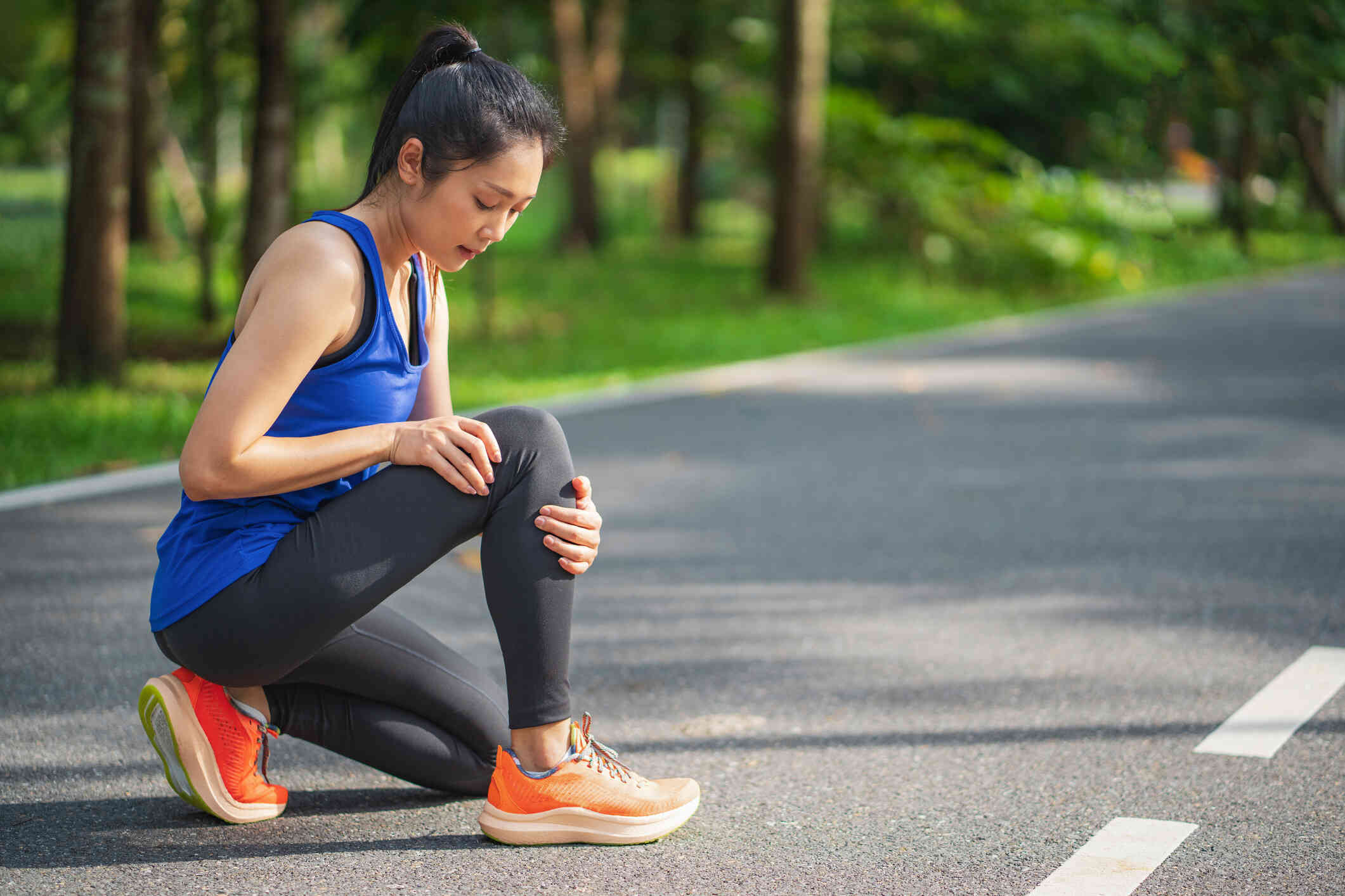 A woman in workout clothes knees down to examine her knee while outside on a walking trail with a serious expression.